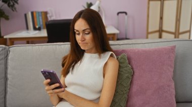 Oturma odasındaki bir kanepede oturan, akıllı telefon kullanan, arka planda kitap rafları ve bavulu olan, beyaz bir elbise giyen ve odaklanmış görünen bir kadın..