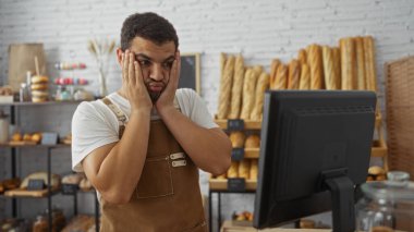 Young man in a bakery shop looking stressed while working on a computer with loaves of bread in the background clipart