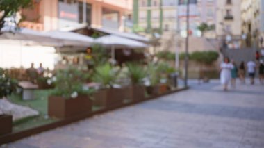 Bulanık insanların yürüdüğü ve oturduğu açık hava kafe sahnesi, kaldırımdaki bitkiler, şehrin arka planı, beyaz elbiseli kadın, sıcak ışıklandırmalı canlı kentsel ortam.