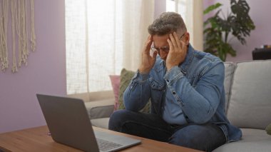 Evde oturan adam stresli görünüyor. Evde dizüstü bilgisayarla çalışırken kafasına dokunuyor.