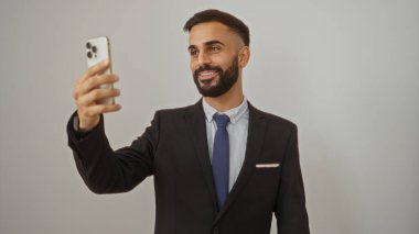Genç İspanyol adam, resmi kıyafetle izole edilmiş beyaz arka plana gülümserken akıllı telefonuyla selfie çekiyor.