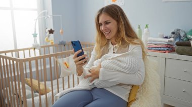 Bebekle gülümseyen bir kadın yatak odasında beşiğinde akıllı telefon kullanıyor, aile içi ve hassas bir anı gözler önüne seriyor, evde genç bir anneliği yakalıyor..