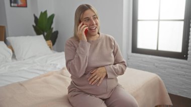 Sarı saçlı hamile bir kadın rahat bir yatak odasında bir yatakta oturuyor, gülümsüyor ve telefonda konuşuyor, elleri göbeğinde, evde rahatlığı ve beklentiyi ön plana çıkarıyor..
