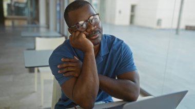 Ofiste oturan, masalar ve sandalyelerle çevrili, mavi gömlekli, düşünceli bir yüzü olan genç bir adam..