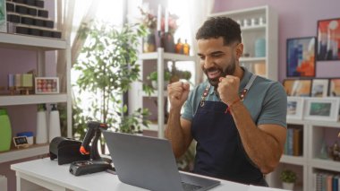 Ev dekorasyonu mağazasında tezgahın arkasında dekorasyon ve bitki dolu raflarla dikilirken dizüstü bilgisayarına tezahürat yapan genç bir İspanyol.