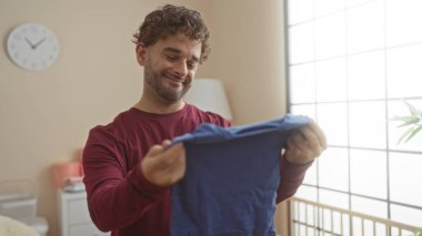 Yatak odasındaki genç adam beşikte bebek kıyafetleri tutarken gülümsüyor, sıcak ve samimi bir ev sahnesi yaratıyor..