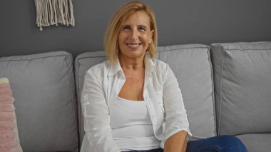 Sarı saçlı, kısa saçlı, gülümseyen olgun bir kadın, evdeki modern oturma odasındaki gri bir koltukta rahatça oturuyor, sıcaklık ve rahatlık saçıyor..