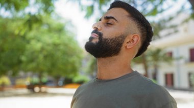 Açık havada sakallı genç bir adam, yeşil ağaçlar ve binalarla dolu bir şehir parkında günün tadını çıkarıyor.