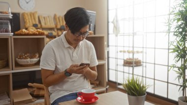Genç Çinli bir adam rahat bir fırında telefon kullanıyor. Yanında taze hamur işleri ve kahve var.