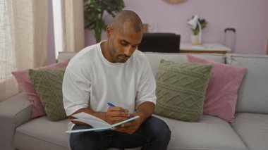 Genç adam, arka planda yastık ve lambayla kanepede otururken deftere bir şeyler yazıyor.