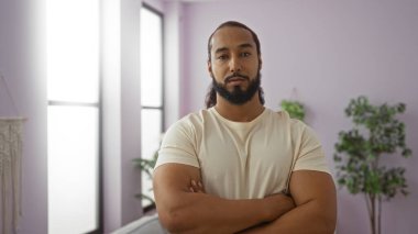 Modern bir oturma odasında kollarını kavuşturmuş sakallı genç bir adam yumuşak ışıklandırma ve şık dekorla sakin bir iç mekan atmosferi sergiliyor..