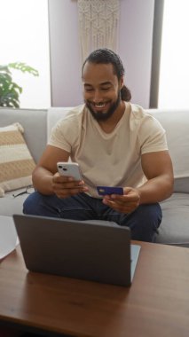 Gülümseyen genç adam, ahşap masada dizüstü bilgisayarla oturma odasında otururken akıllı telefon ve kredi kartını elinde tutuyor..