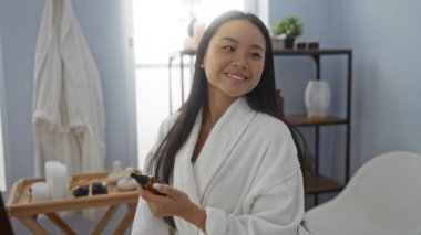 Beyaz bir bornoz giymiş, elinde güzellik ürünleri olan bir kadın spa odasında gülümsüyor. Mumlar ve dekorlu bir raf da dahil..