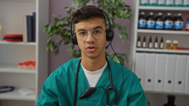 Young man in clinic wearing headset and glasses in a structured room with medical supplies and books visible on shelves behind, conveying a professional work environment. clipart