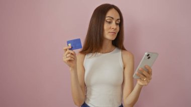 Soyutlanmış pembe arka planda kredi kartı ve akıllı telefon tutan genç bir kadın.