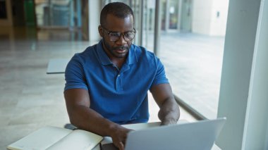 Yakışıklı, genç, Afrikalı, Amerikalı, modern, kapalı bir ofiste dizüstü bilgisayarla çalışan, pencere kenarında oturan, ciddi ve odaklanmış görünen bir adam.