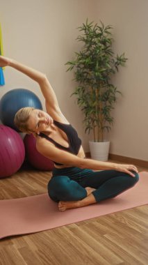 Paspaslı, spor topları ve bitkilerle yoga yapan, huzurlu bir spor salonunda spor yapan, sarışın güzelliğini ve sağlığını vurgulayan bir kadın..