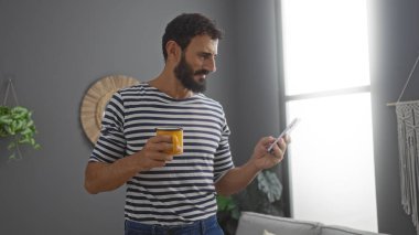 Yakışıklı, sakallı, oturma odasında duran, elinde sarı bir kupayla akıllı telefonuna bakan bir İspanyol..