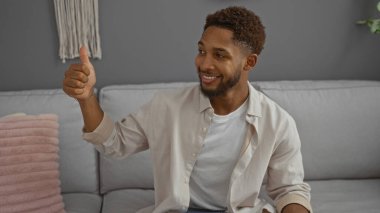 Genç, Afrikalı, Amerikalı, gülümseyen bir adam, modern bir oturma odasındaki bir kanepede oturuyor, pozitif ve rahat bir atmosfer yaratıyor.