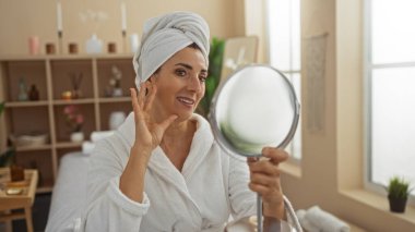 Kafasında havlu olan bir kadın spa odasında ayna tutuyor ve gülümsüyor, bir sağlık merkezinin huzurlu atmosferini yakalıyor..