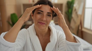 Beyaz bir bornoz giyen, alnına dokunan, kapalı alanda yakalanan, yeşil ve yumuşak ışıkla yeşil ve yumuşak bir çevreyi gösteren, manzaranın güzelliğini artıran bir kadın..