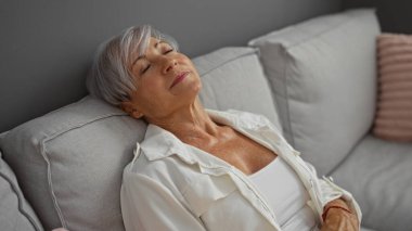 Oturma odasındaki kanepede dinlenen, kısa saçlı, gri saçlı, evde huzurlu bir anın tadını çıkaran bir kadın.