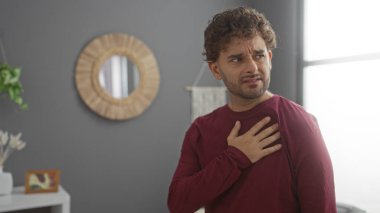 Modern bir oturma odasında elleri göğsünde genç İspanyol bir adam samimi ve şık bir apartman dairesinde duygu ve sıcaklığı resmediyor..