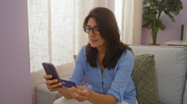 Gözlük takan, elinde kredi kartı olan ve oturma odasında akıllı telefon kullanan bir kadın, arka planda perdeleri olan bir koltukta oturuyor ve odayı süsleyen bitkiler..