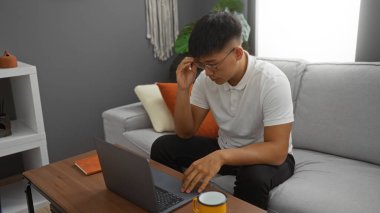 Oturma odasında oturan genç adam dizüstü bilgisayarda çalışıyor ve gözlük takıyor.