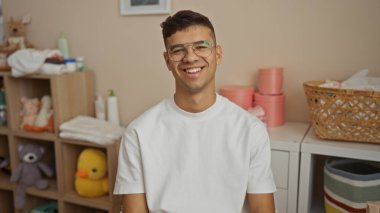 Kapalı bir odada gözlüklü, içi oyuncak ve sepetlerle dolu bir odada gülümseyen genç adam modern bir evde hoş bir ortam yaratıyor..
