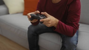 Genç bir adam, rahat bir oturma odasında video oyunları oynuyor, elinde bir kumanda, koltukta oturuyor, rahatlamış, odaklanmış, evde boş vakitlerin tadını çıkarıyor..