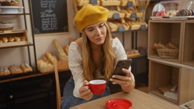 Fırında elinde kırmızı bir bardak ve akıllı telefon tutan genç bir kadın, telefon ekranına bakıyor, sarı bereli ve beyaz bir bluz giyiyor, evin içinde ekmek sergileniyor..