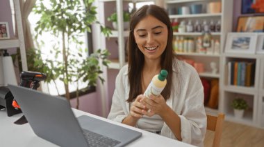Ev dekorasyonları ve bitkilerle çevrili bir mağazada ürün şişesini inceleyen genç bir kadın ve masa başında görünür bir şekilde meşgul ve gülümserken bir dizüstü bilgisayar kullanıyor..