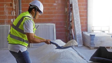 Genç Çinli bir adam kapalı bir inşaat sahasında kumu kürekliyor. Üzerinde sıkı bir şapka ve yüksek görüş mesafesi olan bir yelek var.