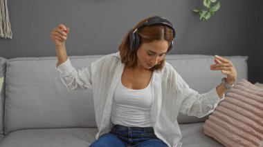 Kulaklıkla müzik dinlerken dans eden genç bir kadın, modern bir oturma odasında bir kanepede oturuyor.