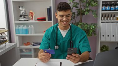 Klinikteki genç adam telefonuna bakıp gülümsüyor ve elinde bir kredi kartı tutuyor. Parlak odada tıbbi eşyalar ve ofis malzemeleriyle çevrili..