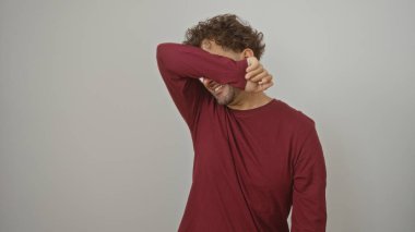 Genç adam koluyla gözlerini kapatıyor, beyaz bir arka plana karşı duruyor, duygularını ifade ediyor..