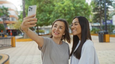 Anne ve kızı şehir parkında selfie çekiyorlar, şehir ortamında bağlarını ve mutluluklarını gösteriyorlar.