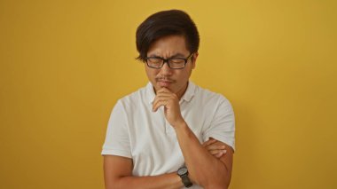 Yakışıklı genç Çinli adam kollarını kavuşturmuş, parlak sarı bir arka plan ve derin düşüncelere dalmış bir ifade karşısında derin derin düşünüyor..