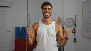 Spor salonundaki spor aletleri ve arka planda beyaz tahtayla duran İspanyol asıllı genç adam, kendine güvenerek gülümsüyor ve fitness merkezli bir ortam gösteriyor..