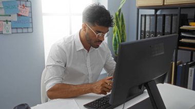 Ofisteki bir bilgisayarda çalışan genç adam görevine odaklanmış. Etrafı dosyalar, dosyalar ve bitkilerle çevrili. Pencereden gelen doğal ışık altında..