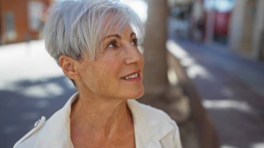 Güzel, gri saçlı, yaşlı, gülümseyen ve yüzünü çeviren bir kadın güneşli, kentsel bir sokakta duruyor..