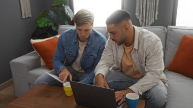Erkek arkadaşlar evlerinde rahat bir oturma odasında dizüstü bilgisayarda çalışıyorlar ve ahşap bir kahve masasında belgeleri tartışıyorlar.