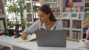 Masasında dizüstü bilgisayar ve diğer ofis malzemeleriyle dekore edilmiş bir mağazada çalışırken bir dergiye notlar yazmaya odaklanan genç bir kadın.