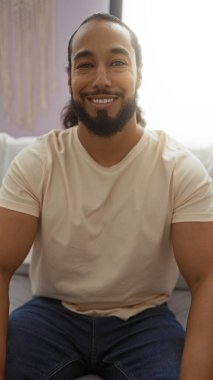 Sakallı genç bir adam modern bir dairenin rahat bir salonunda gülümsüyor, rahat ve yaklaşılabilir bir tavır sergiliyor, yaşam tarzı veya ev dekorasyonu için ideal..