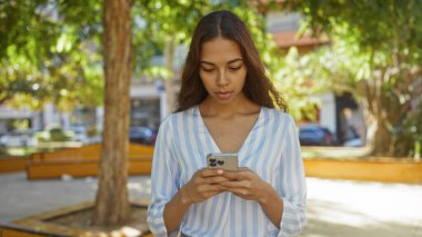 Güneşli bir günde şehir parkında ağaçlar ve şehir binalarıyla çevrili akıllı telefon kullanan İspanyol kadın..