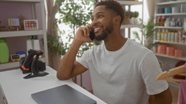 Delikanlı, arka planda renkli eşyalar ve bitkilerle dolu bir mağazada telefonla konuşuyor.