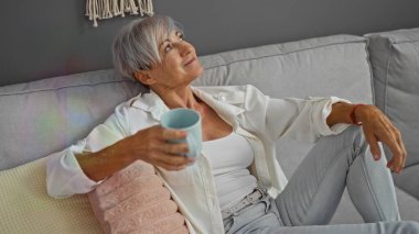 Orta yaşlı bir kadın, kısa gri saçlı, elinde kahve fincanıyla evdeki kanepede dinleniyor ve oturma odasına rahatça bakıyor..