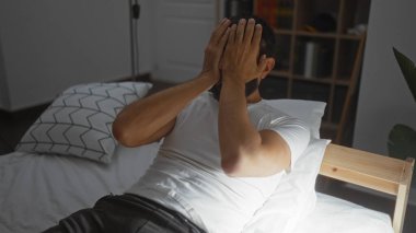 Yatakta sakallı genç bir adam, elleri sıcacık bir yatak odasında yüzünü kapatıyor..