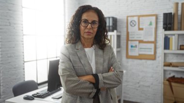 Ofis ortamında kendine güvenen bir kadın, kollarını kavuşturmuş iş kıyafetleri içinde otorite ve zarafeti somutlaştırmış, olgun, İspanyol bir kadını gösteriyor..